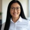 A person with long, straight black hair is wearing a white button-up shirt. The background shows an indoor setting with some greenery and a window, suggesting a professional or office environment.